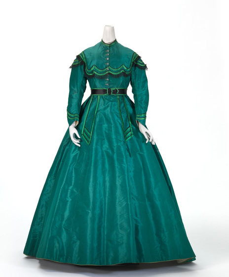 A mannequin wearing a floor length green silk dress, with a lighter green detailing around the wrists, belt line and neck paneling. 
