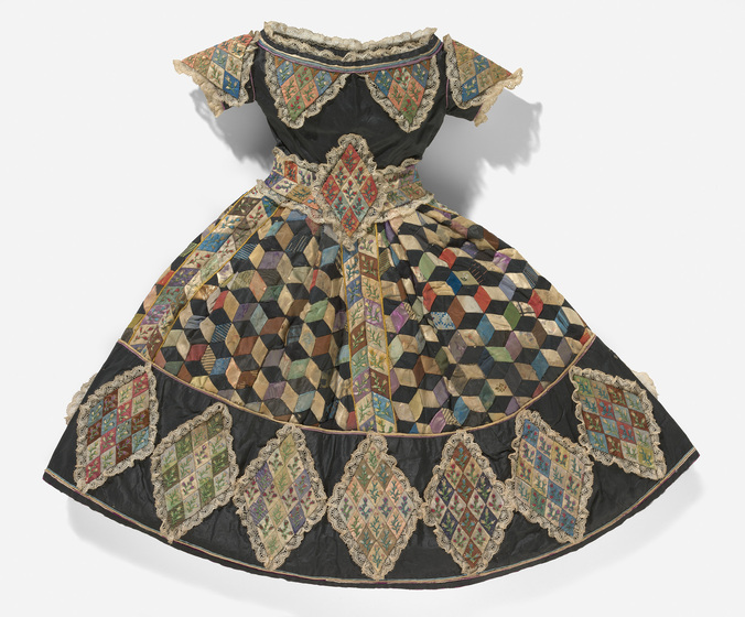 A dolls dress with detailed patchwork patterning around the rim of the skirt, the capped shoulders and the collar, as well as a cinched waist with a diamond shaped belt.