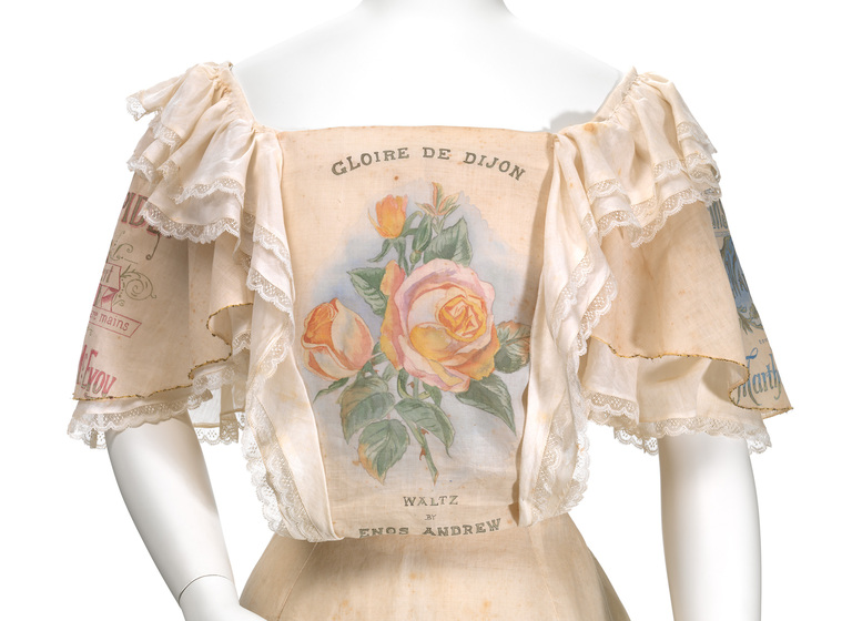 Mannequin wearing a peach coloured top, with loose sleeves to the elbow, and lace featuring on the sleeve ends and shoulder line. The front panel of the top features a printed on bunch of roses, with the words 'Gloire De Dijon'