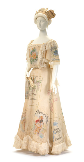 Mannequin wearing peach coloured top and floor length skirt, with lace trim around the bottom of the skirt, the sleeve cuffs, shoulder lines and down the front of the top. On the clothing are decorative prints of people, scenes and writing in varying colours and fonts. The mannequin is wearing a bonnet with sheets of music, positioned around the head line.