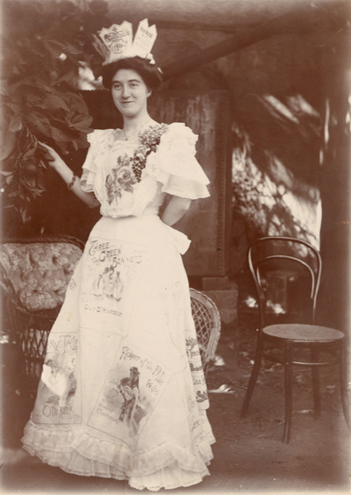 Black and white photograph of a woman wearing a floor length skirt and top featuring various drawings and lettering in different fonts and sizes. She is wearing a crown shape hate with sheet music attached to the rim. She is holding the leaves of a nearby tree, whilst two chairs can be seen behind her.