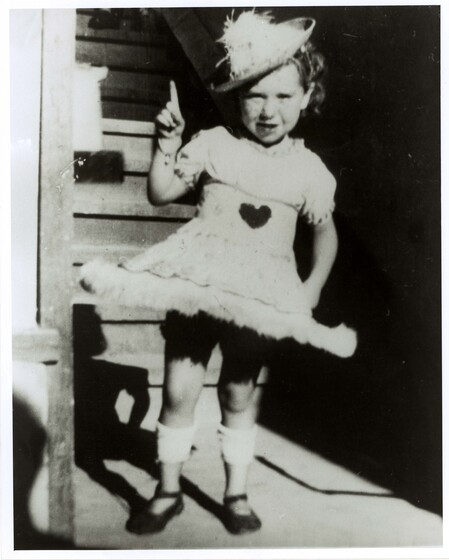 Small girl stands in a short dress with a hoop skirt, knee high socks and a hat with feathers on top. She has one hand on her hip, and hand hand in the air with her finger pointing up.