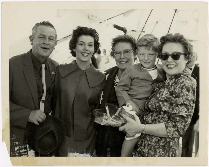 A group of four adults, three female and one male stand together in a group smiling at the camera. One of the women is holding a small boy on her hip, his arm wrapped around her neck.