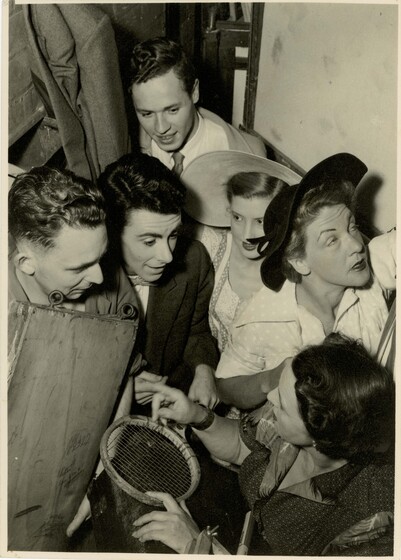 A group of adults stand in a small huddled group, each of them looking in different directions. One of them is holding a small faced tennis racket.
