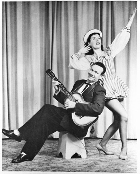A man sits on a stool, his legs crossed over, playing a guitar and looking at the camera. Behind him stands a woman in a short dress and hat, striking a pose with one hand behind her ear and one arm in the air. They're standing behind a floor length curtain.