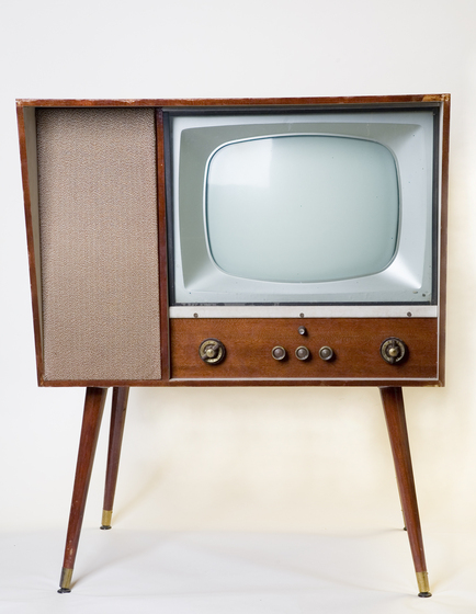 A square framed tv made from wooden paneling, and four wooden legs. The television screen is positioned to the left, with brass dials positioned below it.