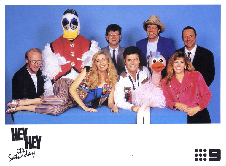 A group of men and women stand around a blue box. On the blue box is a woman lying down and a pink ostrich puppet. In the back row is a person dressed in a duck suit, wearing a red vest and a baseball hat.