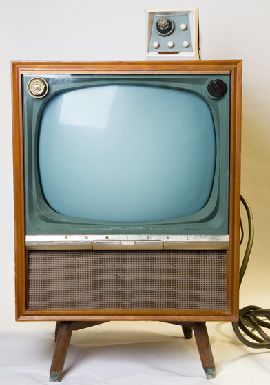 A television set framed within a wooden box, positioned on four wooden legs. A large speaker area sits below the screen, and a smaller box is attached to the top, featuring dials of different sizes.