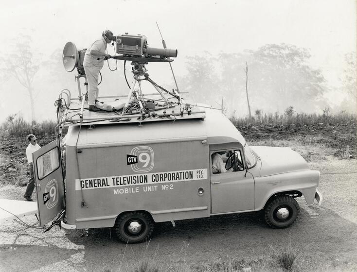 Photograph of a van parked on the side of a road with landscape around. On top of the car stands a man looking through a film camera, maneuvering it with handles. The back door is open and a man can be seen walking past the back entry of the van.
