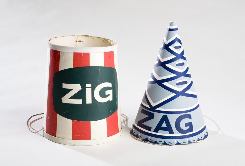 Two paper hands, one rounded at the top and one pointy/ The rounded hat has red and white stripes with a green label on the front saying 'Zig'. The pointed hat has blue and white criss crossing stripes with lettering on the front saying 'Zag'.