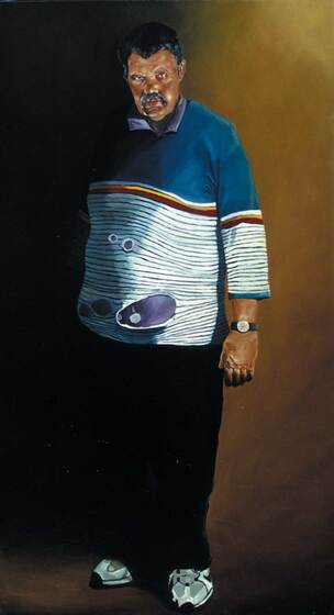 Portrait of a man standing, leaning slightly against a wall. He has a very serious look on his face and is dressed in a striped jumper with a blue section on top, black pants and white sneakers.