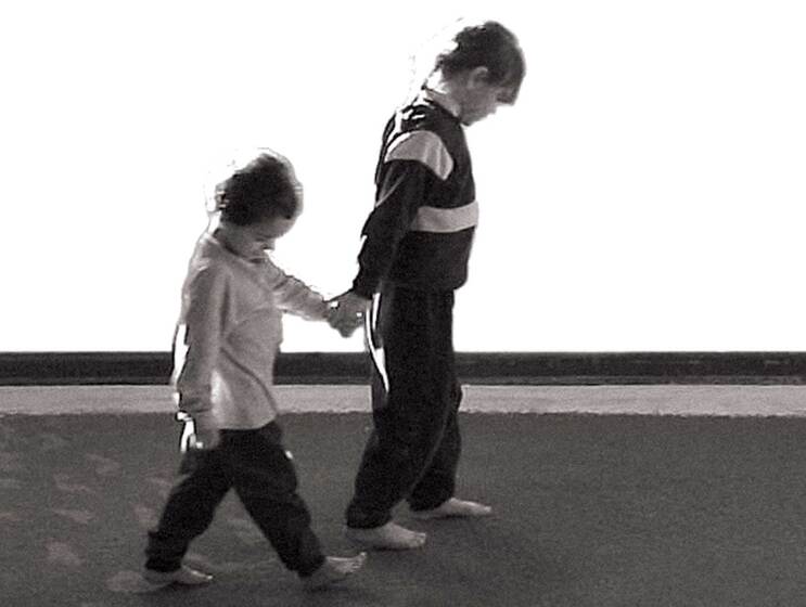 A black and white photograph of two children walking hand in hand, both looking down at the ground, they're leaving footprints on the ground behind them.