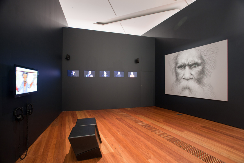 Exhibition space showing three black walls, one with a large portrait of William Barak, one with 5 small screens, and one with a medium sized screen and headphones hanging below. Two stools are positioned in the middle of the room.