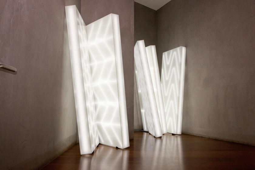 Two large rectangular white light installations lean against a concrete wall. They have wave like patterns across the surface, appearing slightly brighter than the rest of the space.