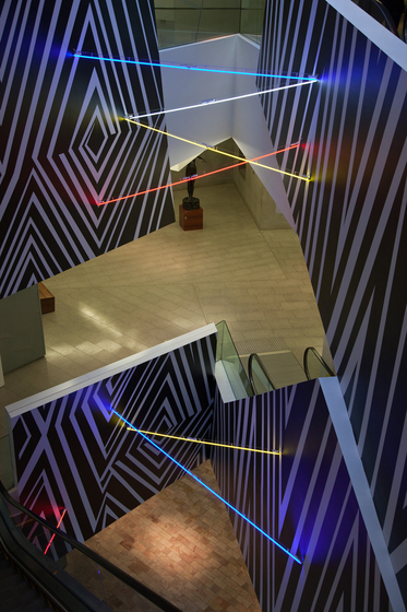 A photograph of an exhibition space, looking from a high up level of a building, down to the two levels below. On the walls of the lower levels are black and white patterns of vortexes and lines, with neon light bars criss-crossing between the open spaces.