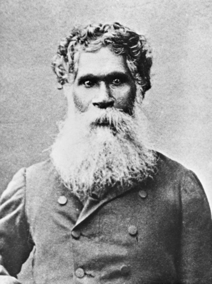 Portrait photograph of an indigenous man staring directly at the camera. He is wearing a buttoned up woolen coat and has a long white beard covering his chin and neck.