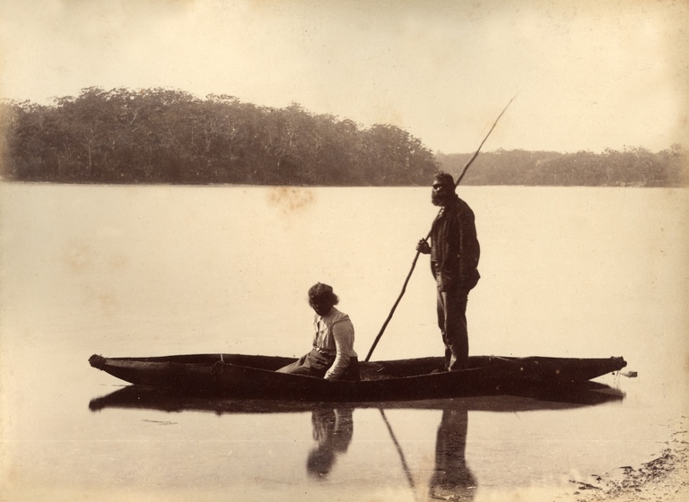 A man and a woman in a bark canoe on a serene lake, trees lining the distant shore behind them. The man stands up in the canoe with a spear held over his back shoulder, whilst the woman sits in the front of the canoe, her face turned to the camera. They are both wearing European clothing.