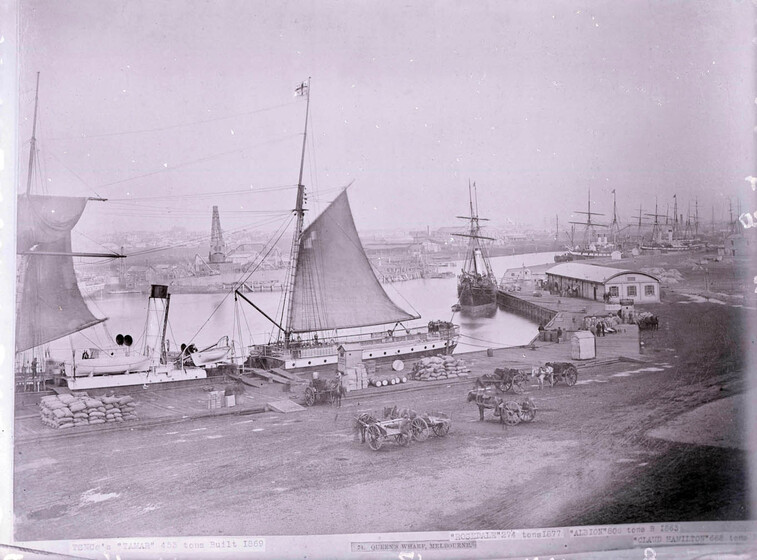 Black and white photograph of a shipping wharf, with multiple sailing ships docked along the edge, stretching into the distance. In the foreground on the wharf, horse and carts stand with goods, and piles of sand bags are stacked in groups.