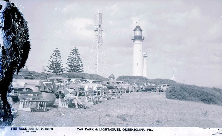 Black and white postcard of a large group of cars, all lined up next to each other and facing the waters edge. In the background is a white lighthouse and telegraph tower, with green shrubs framing the scene.