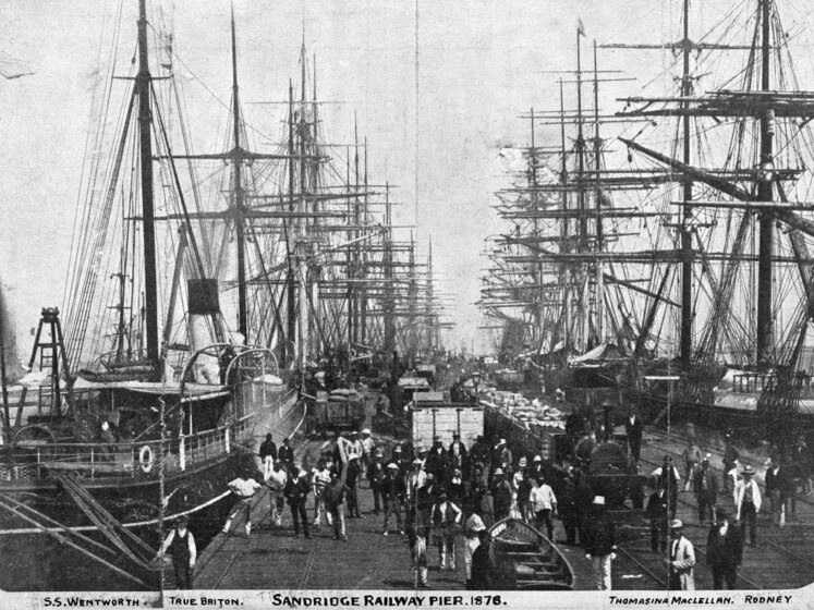 Photograph of a busy pier, with tall ships lined either side, moored and awaiting stocking. Along the wharf itself, men are at work moving materials. A small train can be seen, its carriages piled high with contents.