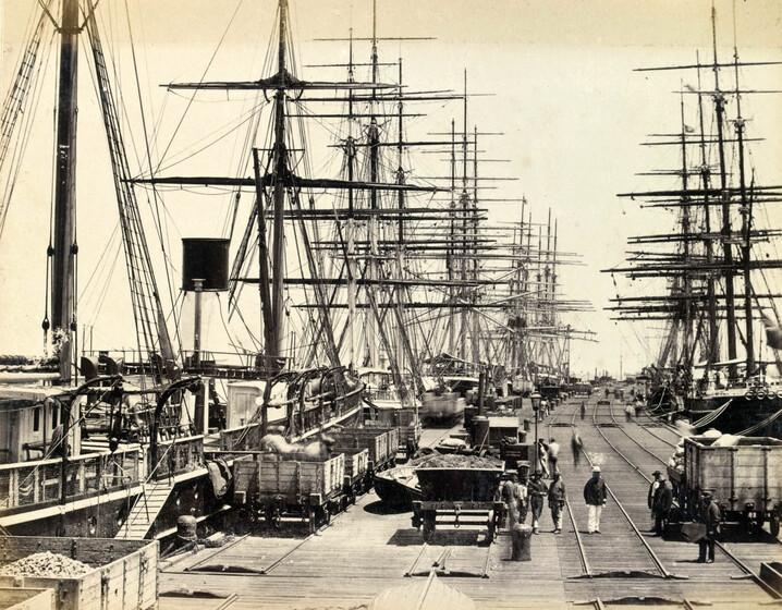 Photograph of a busy pier, with tall ships lined either side, moored and awaiting stocking. Along the wharf itself, men are at work moving materials. Railway carriages line the piers edge, some full of materials, and others waiting to be filled. Railway tracks stretch down the pier into the distance.