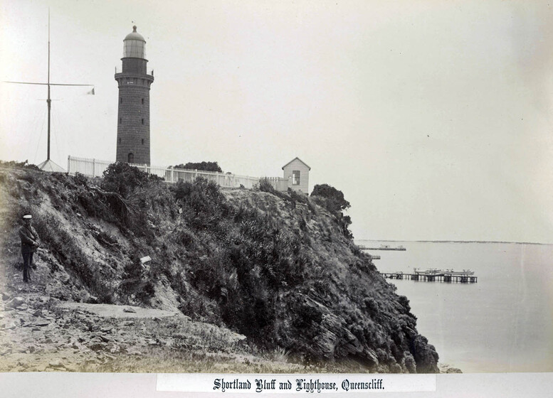 Photograph of a small lighthouse and cottage, perched on a rocky outcrop. In the foreground, a man stands on the edge of the cliff looking at the ocean. In the distance, small jetty's stretch out into the water.