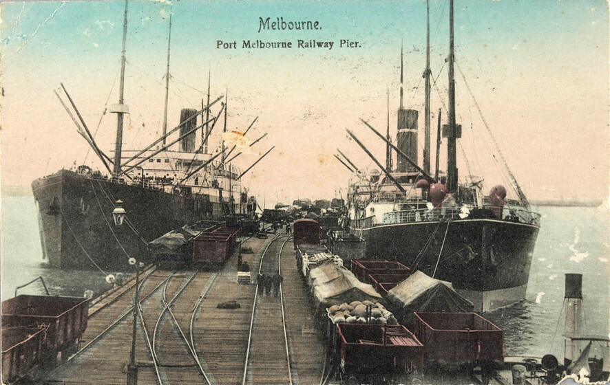 Two large steam boats are docked either side of a wide wooden pier. Along the pier run railway tracks, including some that are being use by red wagons full of materials.