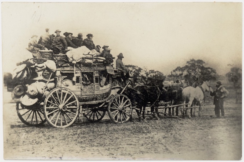 Sepia photograph of a horse and cart, laden with bags and materials, and on top a group of Chinese men. At the front of the wagon, in front of the 6 horses stands a man feeding the front horse.
