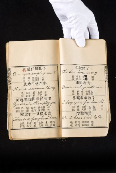 Double page book with Cantonese characters and English translations, being held by a hand with a white glove on.