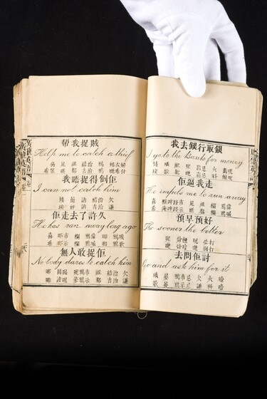 Double page book with Cantonese characters and English translations, being held by a hand with a white glove on.