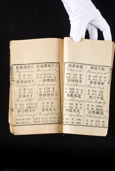 Double page book with Cantonese characters and English translations, being held by a hand with a white glove on.