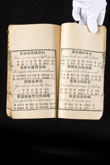 Double page book with Cantonese characters and English translations, being held by a hand with a white glove on.