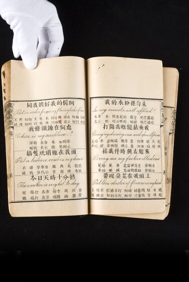 Double page book with Cantonese characters and English translations, being held by a hand with a white glove on.