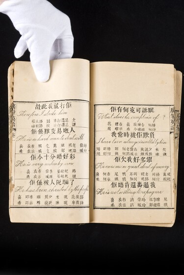 Double page book with Cantonese characters and English translations, being held by a hand with a white glove on.
