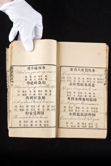 Double page book with Cantonese characters and English translations, being held by a hand with a white glove on.