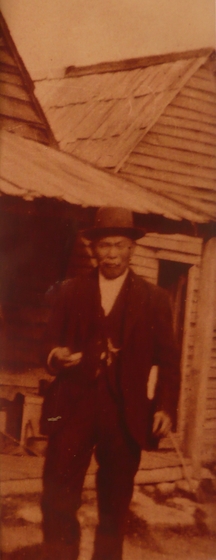 An older Chinese man in a three piece suit, standing in front of a weatherboard house.