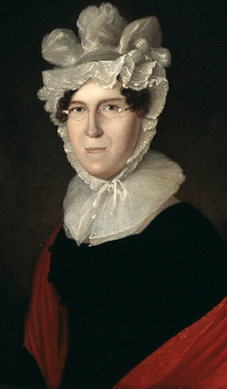 Portrait of a middle aged woman dressed in a decorative white bonnet, wire rimmed glasses, a black dress and red cloak, resting partway down her shoulders.