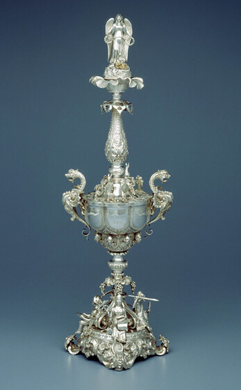 A decorative standing cup, covered in highly decorative silver adornments, inclyding women playing instruments, dragons and other decorative patterns. On the top of the cup is an angel with full length wings and arms stretched out.