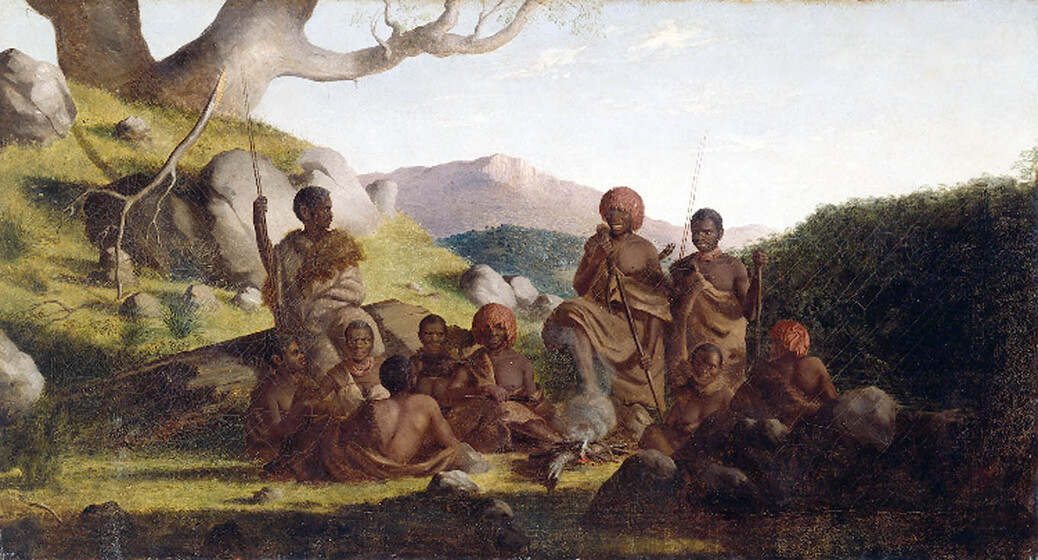 A group of indigenous men and women stand and sit around a small campfire. There are large boulders nearby, and a large tree slightly in the distance. Some of the group are wearing robes and head adornments.