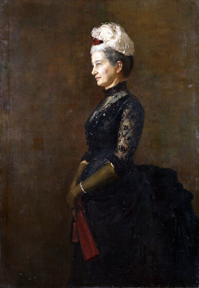 Portrait of a well dressed woman in a full length black dress, the bustle sticking out someway from her waistline. The sleeves are made of decorative black lace, and she is wearing gold gloves and a white feathered hairpiece.