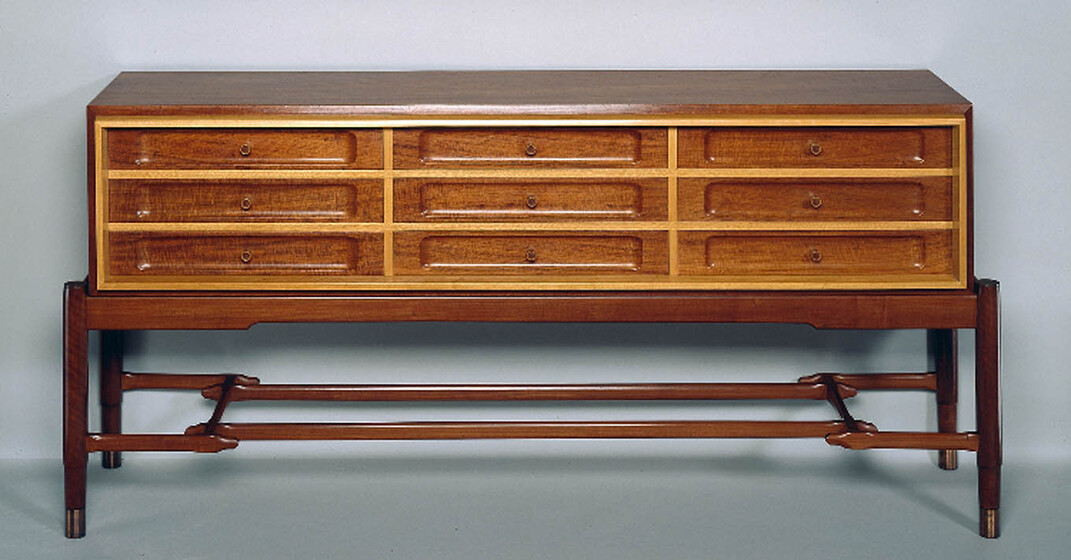 A light brown shallow chest standing on a wooden frame made of rounded legs and cross frame. The chest itself has 9 drawers on the front in a 3 by 3 pattern.