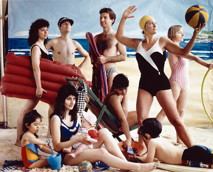 A posed photograph of a group of adults and children standing and sitting on a makeshift beach. A backdrop canvas of the ocean and sand is positioned behind the group. Each of the individuals is in some kind of bathing outfit, with many holding props like buckets, towels, lilo's and balls.