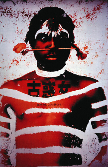 An indigenous man stands topless with white markings across his chest and arms. He has chinese characters printed on his chest, a bar through his nose and some kind of head piece made of bones.