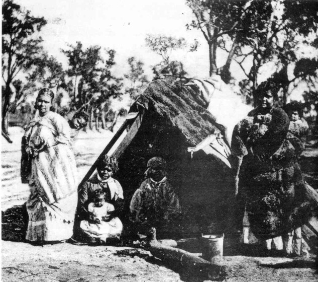 Black and white photograph of a group of indigenous people standing and sitting in front of a bark hut. Some of the women are wearing possum skin cloaks, each with babies strapped to their back.