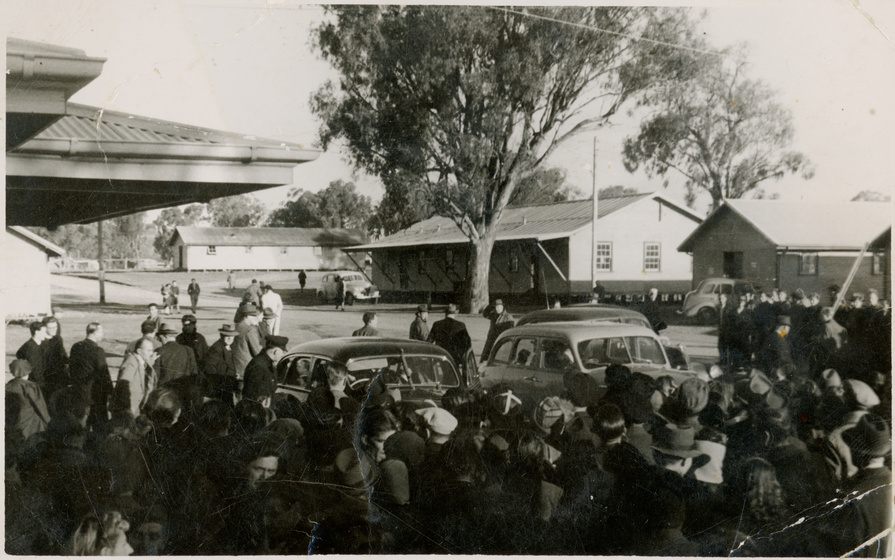 Black and white photograph from the 1950s or 60s of a moderate crowd of people standing around three parked cars. Behind the group of people are wooden buildings and large gum trees.