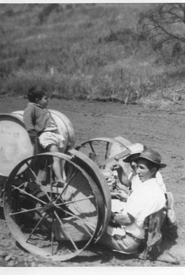 Black and white photograph of two men sitting on the ground with large iron wheels positioned either side of them. A small child leans on the farm machinery, staring into the distance.