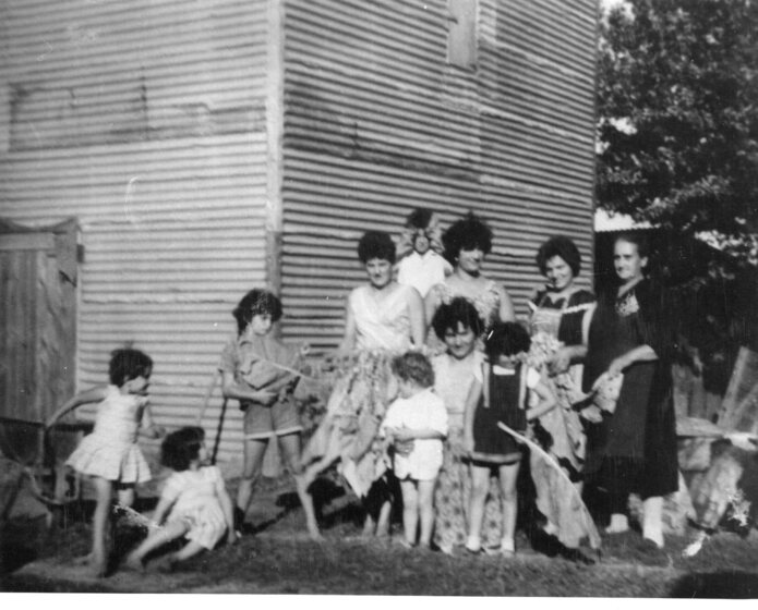 A group of children and women stand in front of a corrugated iron tobacco kiln.