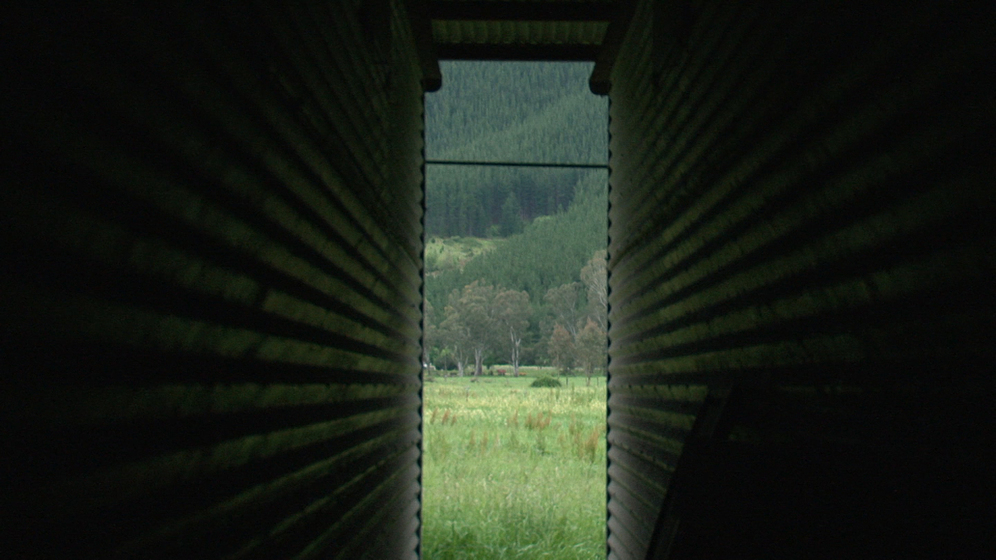 Photograph taken between the gap of two tin sheds, looking out onto a slither of green paddocks and tree lined hills.