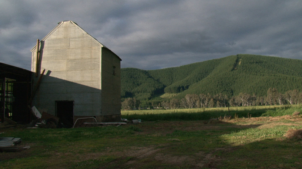 A corrugated iron tobacco kiln stands to the left, slightly falling down and rubble around the ground. Behind the kiln are tree covered hillsides and a moody grey sky above.