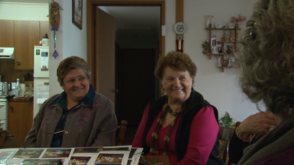 Two older women sit at table in a home setting - they are both smiling and looking at a third person who is partially in the shot, their head turned away from the camera. On the table is a photo album, opened with photographs showing.